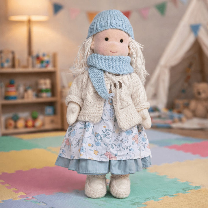 Handmade Waldorf-style cotton doll with blonde hair, a blue knit hat and scarf and a floral skirt, standing in a playroom