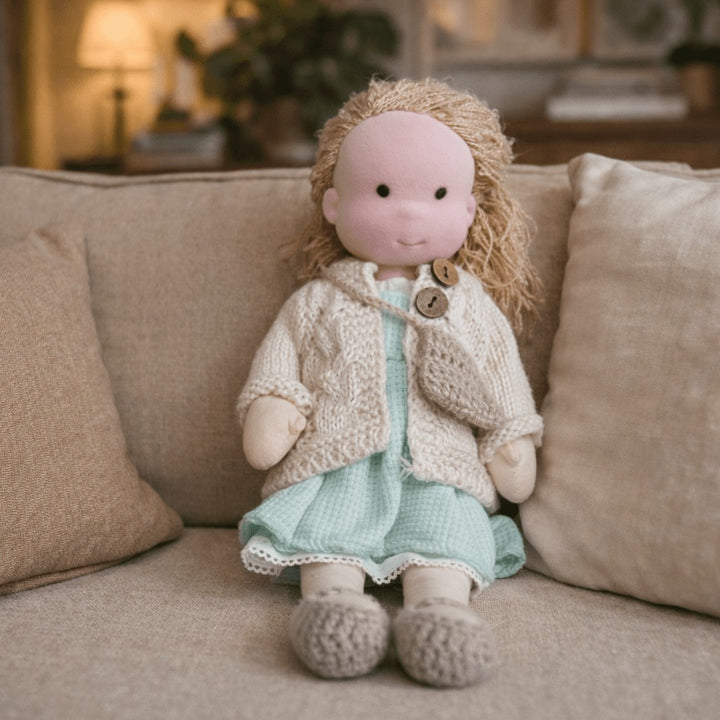 Handmade Waldorf-style cotton doll with blonde curls, a mint dress and a cream knit cardigan seated on a sofa