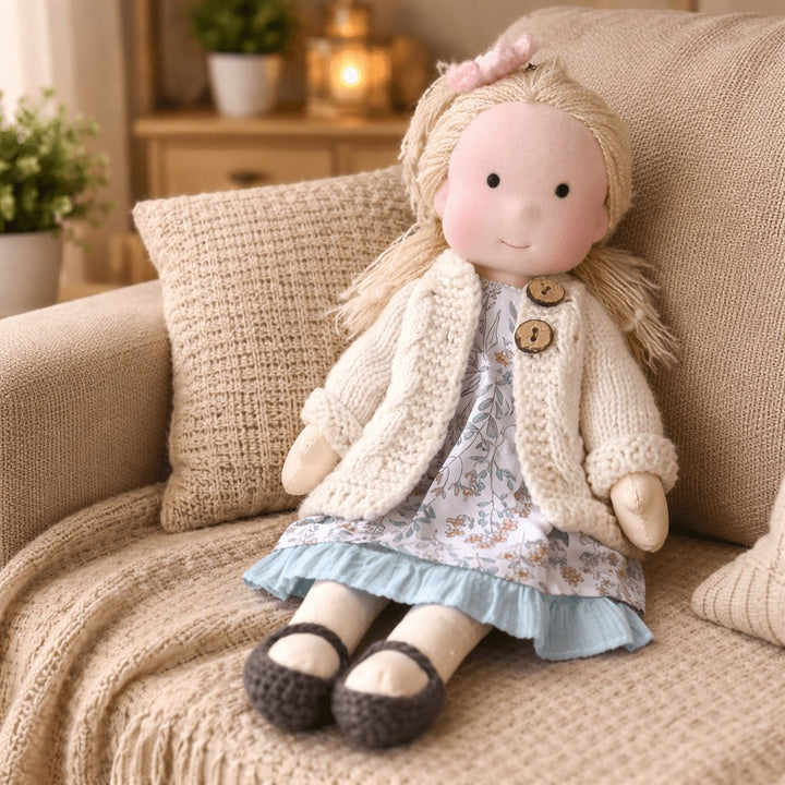 Handmade Waldorf-style cotton doll with blonde hair, a cream knit cardigan and a pale floral skirt on a sofa