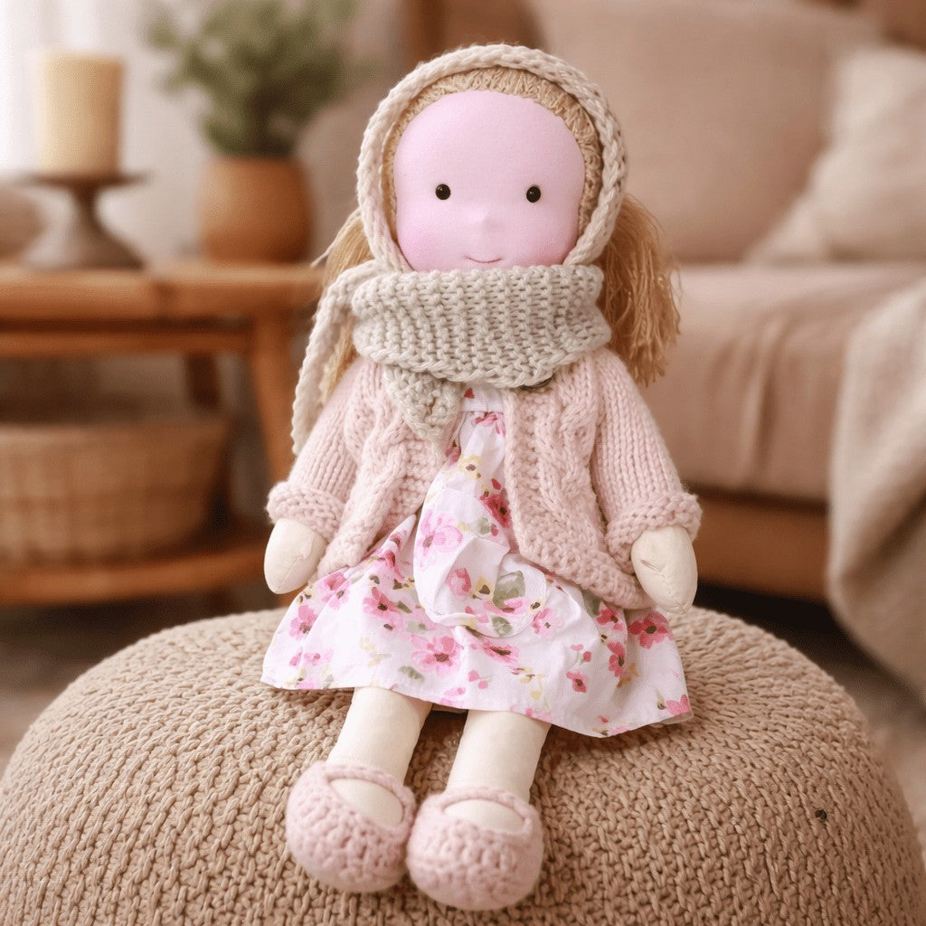 Handmade Waldorf-style cotton doll with a cream bonnet and scarf and a pink floral dress on a knit pouf