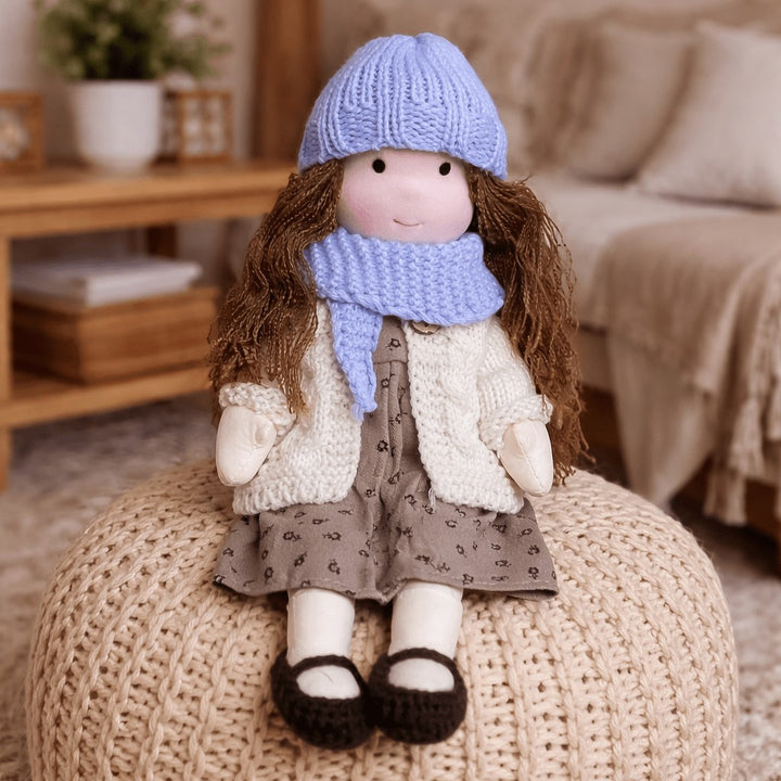Handmade Waldorf-style cotton doll with brown hair, a blue knit hat, lilac scarf and a brown floral skirt on a knit pouf