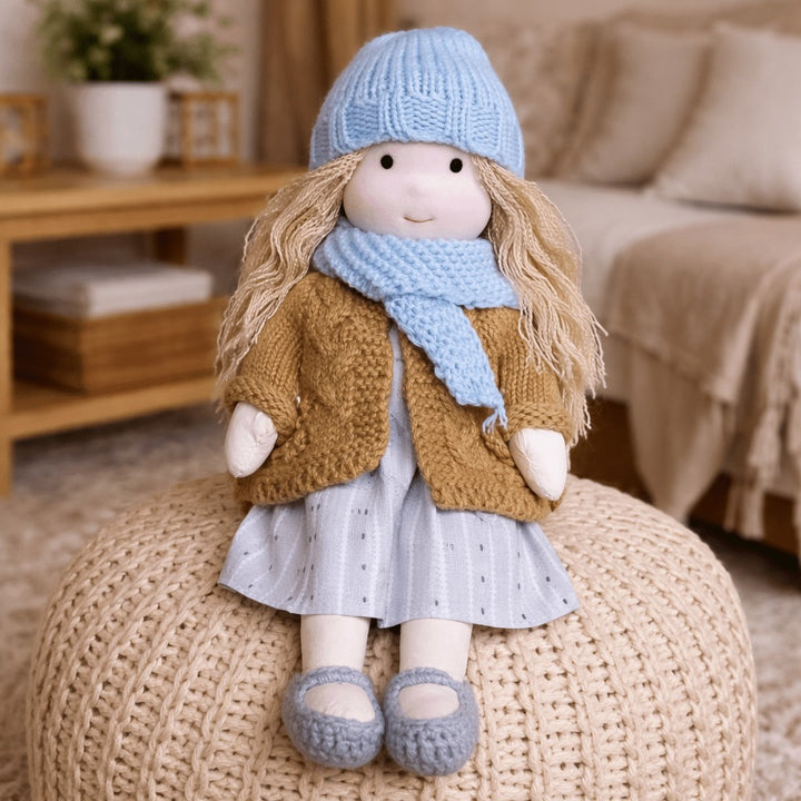Handmade Waldorf-style cotton doll with blonde hair, a blue knit hat and scarf and a mustard cardigan on a knit pouf