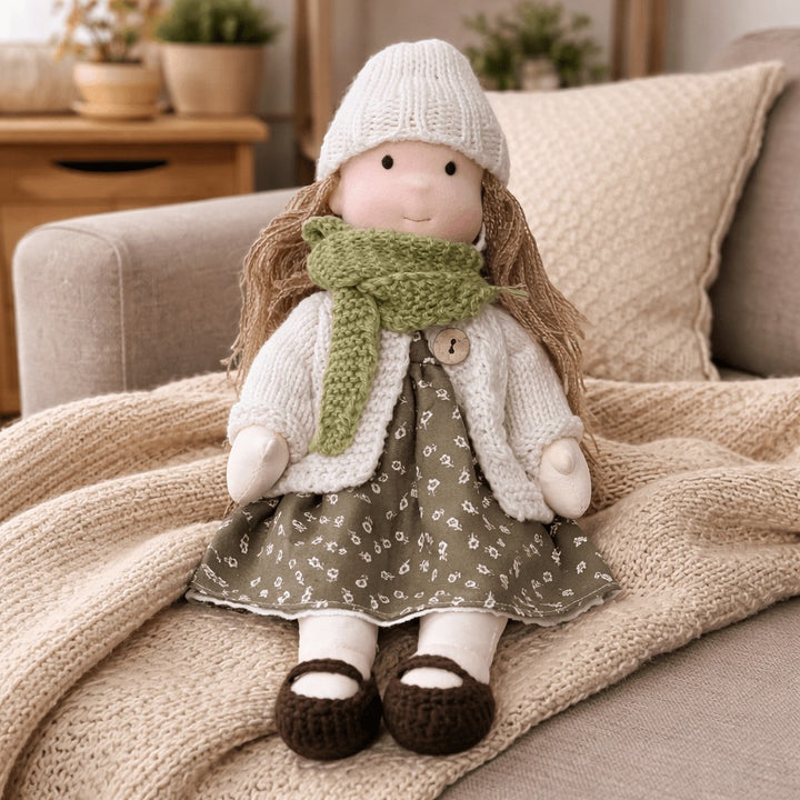 Handmade Waldorf-style cotton doll with brown hair, a cream knit hat, green scarf and an olive floral skirt on a blanket